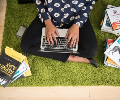 tyttö istuu lattialla kannettava tietokone sylissään kirjojen ympäröimänä